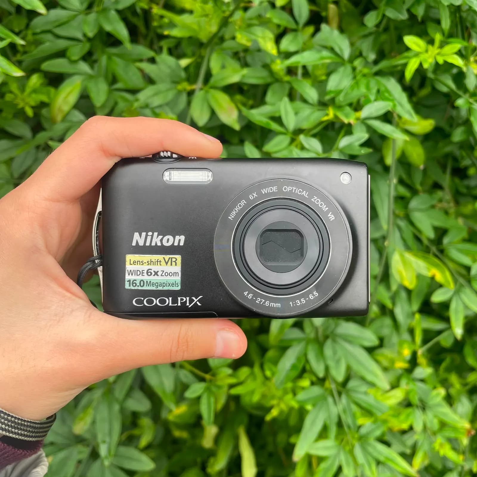 Cámara Nikon Coolpix negra sostenida en una mano, de frente, con el objetivo NIKKOR de 6 aumentos